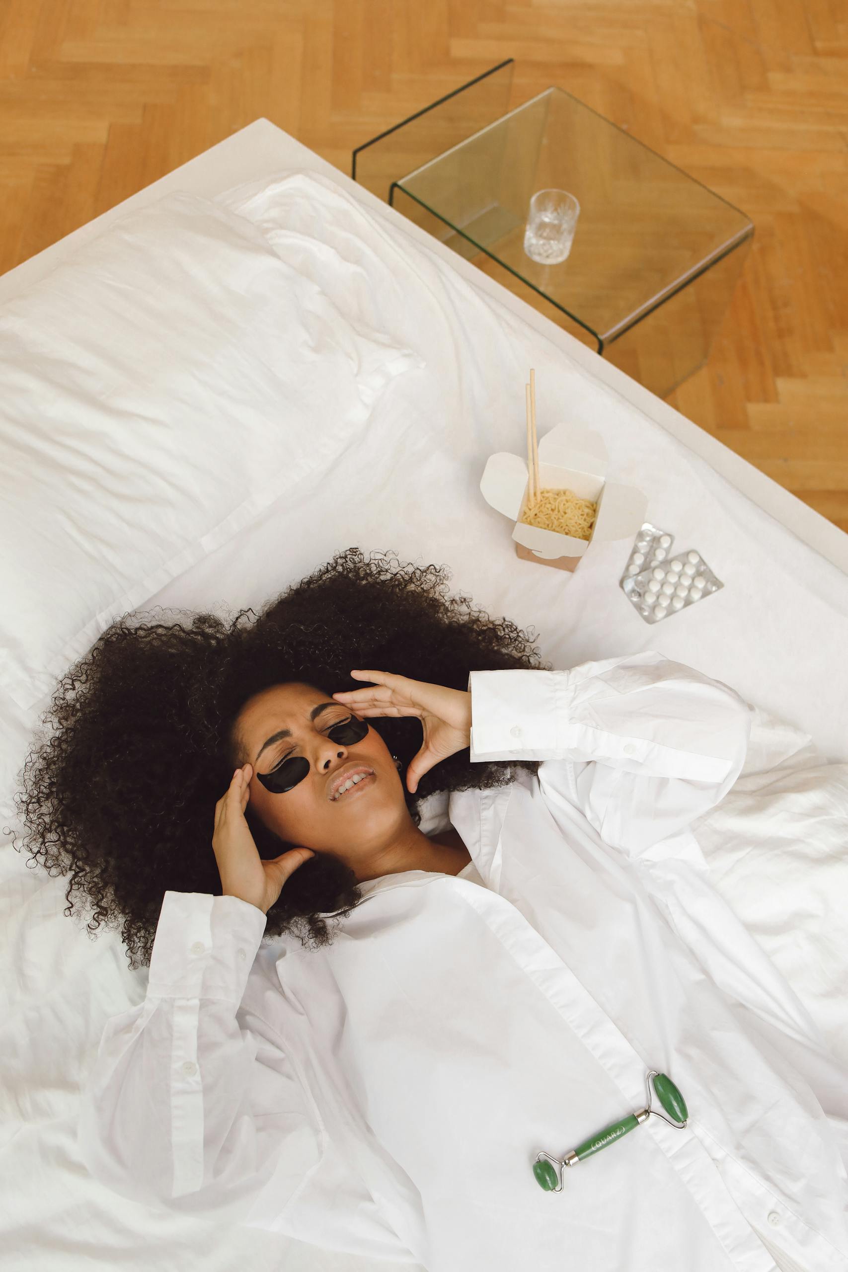A woman in a white dress shirt lying on bed with jade roller and under-eye masks, expressing relaxation.